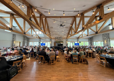 Large banquet hall with exposed wooden beams and warm string lights, rows of tables filled with guests dining.