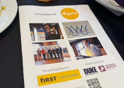 Program flyer on a dark table with 'Celebrate Aspire' logo, sponsor images, and a nearby chocolate chip cookie; a person in purple sleeve reaches toward a plate.