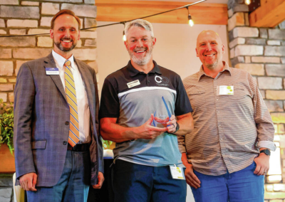Three men standing indoors at a ceremony; center man holds a glass award and smiles, with two colleagues on either side.
