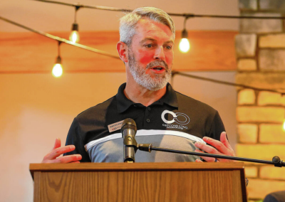 Man with gray hair and beard speaks at a wooden podium with a microphone, wearing a black polo with a logo.